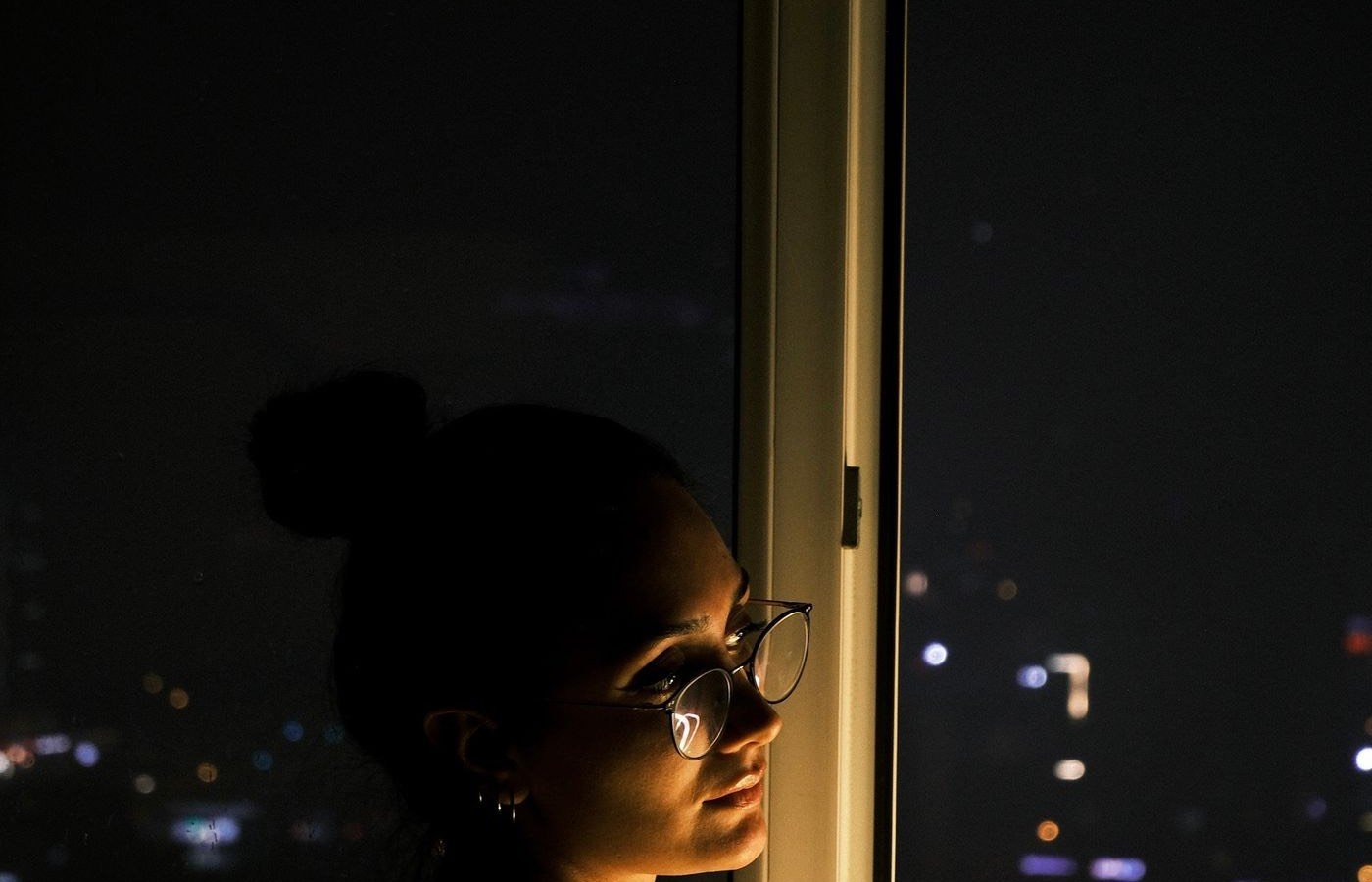 Una mujer sola frente a la ventana de noche, mirando las luces de la ciudad